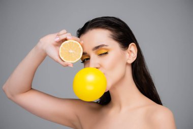 Makyajlı genç esmer kadın sarı sakız üflüyor ve limonun yarısını gri renkte soyutluyor.