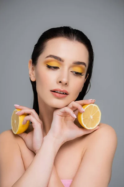 brunette woman with bare shoulders holding yellow lemon halves isolated on grey
