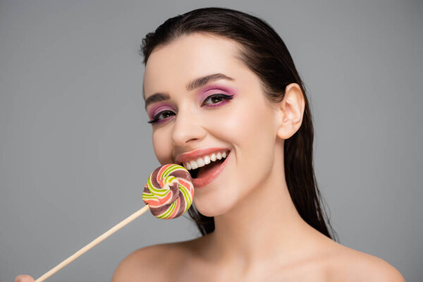 smiling young woman with pink eye shadows holding lollipop isolated on grey 