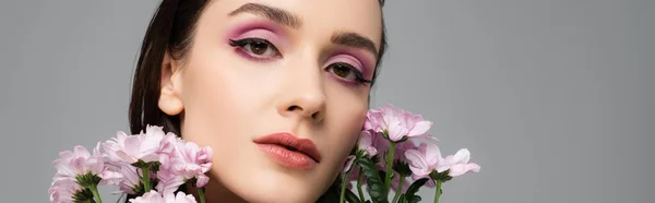 young woman with pink eye shadows near flowers isolated on grey, banner