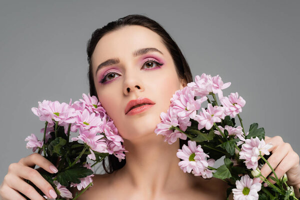 young woman with pink eye shadows holding flowers and looking up isolated on grey 