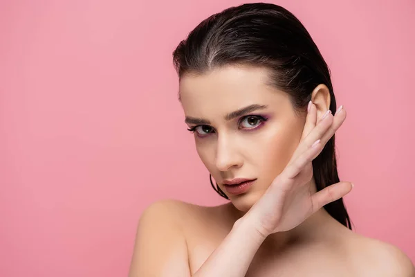 sensual woman with makeup and bare shoulders looking at camera isolated on pink