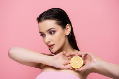 Çıplak omuzlu genç bir kadın olgun limonun yarısını tutuyor ve pembelerden soyutlanmış bir şekilde uzaklara bakıyor.