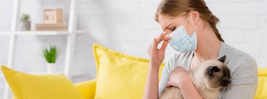 Woman in medial mask suffering from allergy reaction and holding siamese cat, banner 