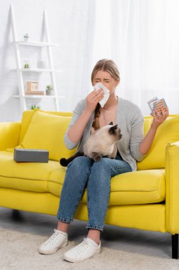 Woman with allergy sneezing while holding pills and siamese cat  clipart