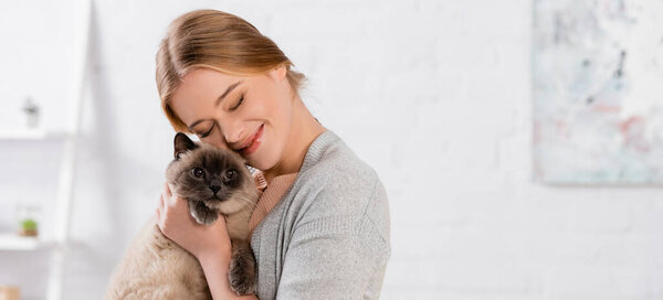 Cheerful woman with closed eyes embracing siamese cat, banner
