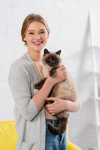 Happy woman looking at camera while holding siamese cat at home 