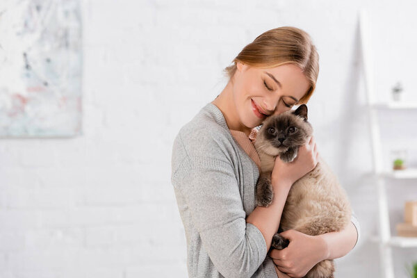 Smiling woman with closed eyes embracing siamese cat 