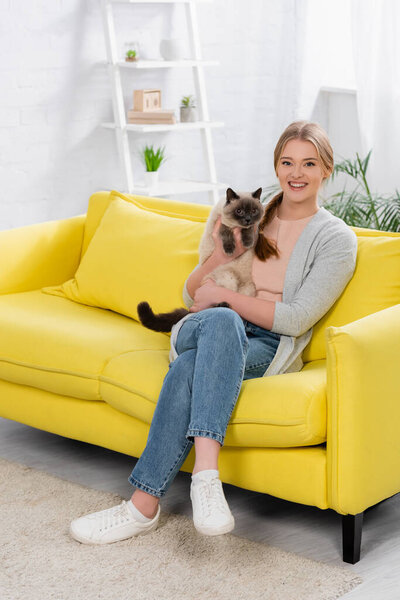 Woman in casual clothes smiling while holding siamese cat on couch 