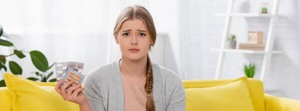 Sad woman with allergy holding blisters with pills and looking at camera a home, banner 