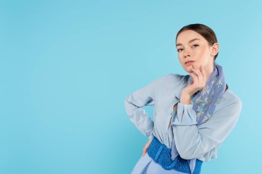 Yüze dokunan ve kameraya bakan şık bir kadın elleri kalçasında mavinin üzerinde dikilirken.