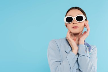 Şık güneş gözlüklü çekici bir kadın. Yüzü maviye yakın pozlarda poz veriyor.