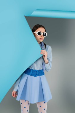 Gri arka planda mavi yırtık kağıt yanında moda elbiseli ve güneş gözlüklü genç kadın.