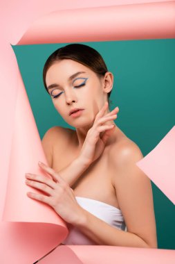 Kapalı gözlü genç bir kadın yeşil kağıttaki deliğin yanında yüzüne dokunuyor.