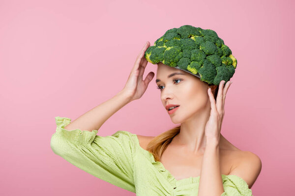 young woman adjusting hat made of broccoli while posing isolated on pink, surrealism concept