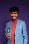 gepiercter afrikanisch-amerikanischer Mann im blauen Blazer lächelt, während er ein Glas Champagner auf lila hält