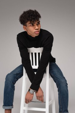 Balıkçı yaka kazak giyen kıvırcık saçlı Amerikan modeli gri renkte poz verirken sandalyede oturuyor.