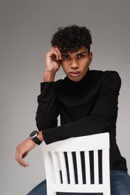 Balıkçı yaka kazaklı Afro-Amerikan adam. Gri sandalyede beyaz sandalyeye yaslanıyor.