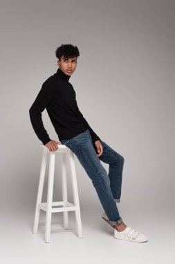 full length of african american young man in turtleneck sweater leaning on white chair on grey clipart