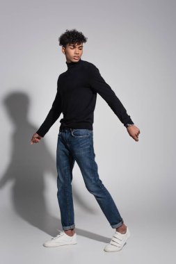 Siyah balıkçı yaka kazaklı, gri üzerinde yürüyen kıvırcık Afrikalı Amerikalı bir adam.