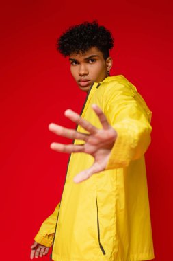 stylish african american man in yellow rain jacket showing stop gesture isolated on red