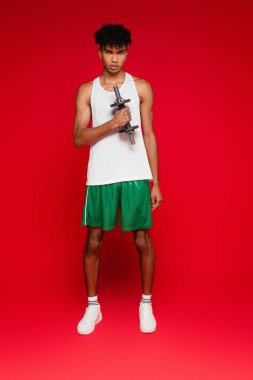 Uzun boylu, şortlu ve kolsuz tişörtlü Afro-Amerikan bir adam. Kırmızı halterli halterle egzersiz yapıyor.