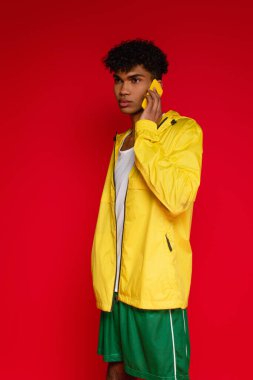 Kıvırcık Afrikalı Amerikalı ceketli ve şortlu adam kırmızıda izole edilmiş akıllı telefondan konuşuyor.