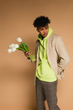young african american man in green hoodie and blazer holding tulips on beige clipart