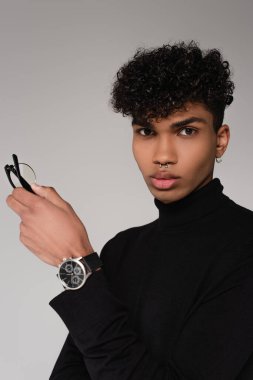 Siyah balıkçı yaka kazak giyen, gözlüklü ve gri üzerine izole edilmiş poz veren delinmiş Afro-Amerikan bir adam.