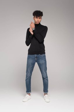 Siyahi balıkçı yaka kazak ve kot pantolonlu, gri üzerine poz veren Afro-Amerikan bir adam.