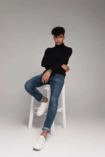 full length of young african american man in turtleneck sweater and jeans sitting on chair while posing on grey