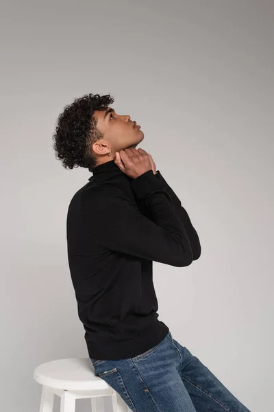 side view of african american man sitting on chair and adjusting collar of turtleneck sweater isolated on grey