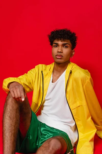 Sarı ceketli kıvırcık Afrikalı Amerikalı adam kırmızı üzerine oturmuş poz veriyor.