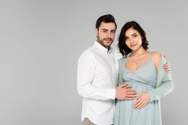 Beyaz tişörtlü, gülümseyen adam. Gri elbiseli hamile eşi kucaklıyor. 