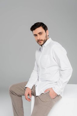 Şık bir adam pantolonun cebinde el ele tutuşup gri arka plandaki küpteki kameraya bakıyor.