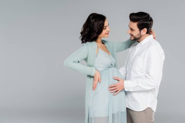 Gülümseyen adam, gri elbiseli hamile kadının karnına dokunuyor. 