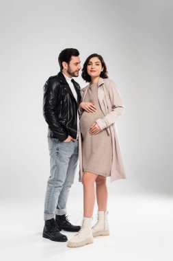 Gri arka planda, kocasının yakınındaki kameraya bakan son moda hamile kadın.