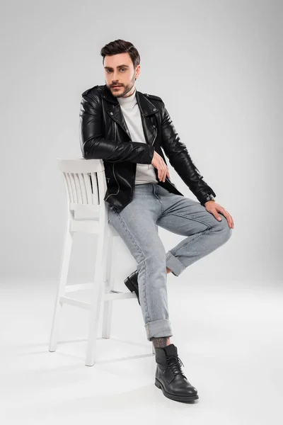 Stylish man in leather jacket looking at camera on chair on grey background