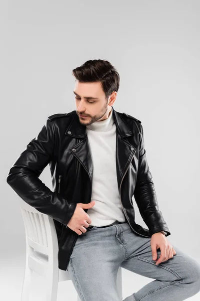 Brunette man in leather jacket sitting on chair isolated on grey