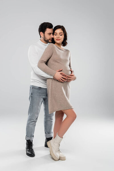 Stylish man hugging smiling pregnant woman in dress and boots on grey background 