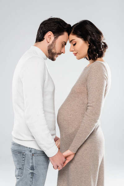 Side view of pregnant woman and husband holding hands isolated on grey 