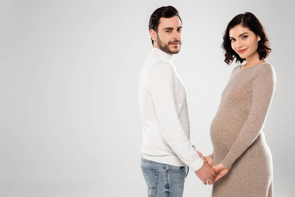 Smiling pregnant woman holding hand of bearded husband isolated on grey 