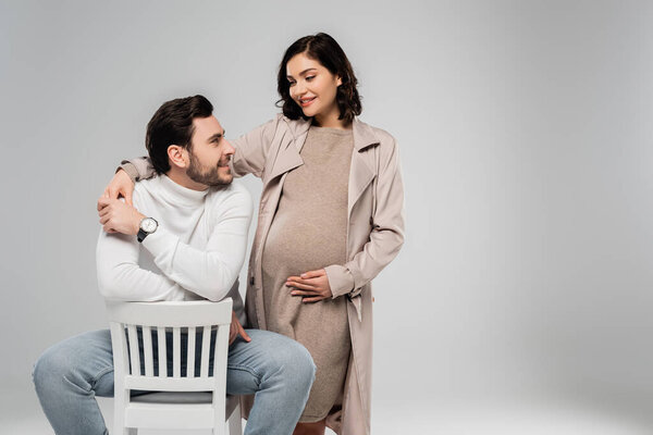 Cheerful pregnant woman looking at husband on chair isolated on grey 