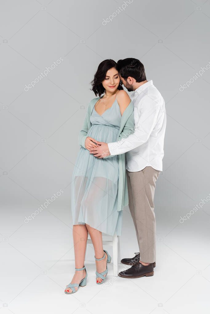 Man hugging pregnant wife near white chair on grey background