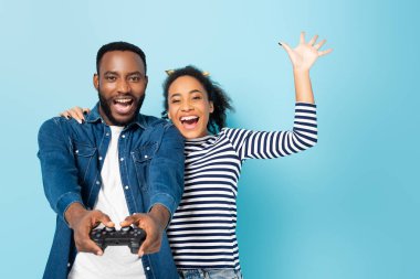 KYIV, UKRAINE - OCTOBER 23, 2020: excited african american woman waving hand while hugging husband gaming on blue clipart