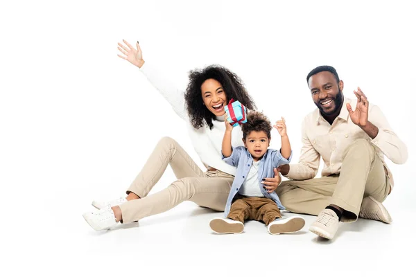 stock image african american boy holding gift box near cheerful parents waving hands on white