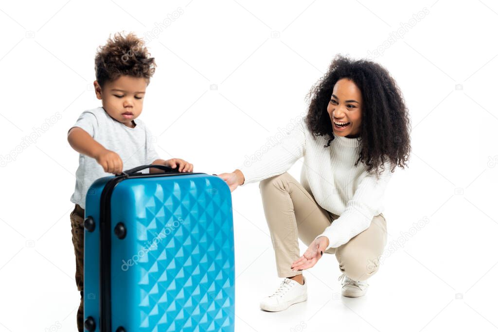 laughing african american woman pointing at travel bag near little son on white