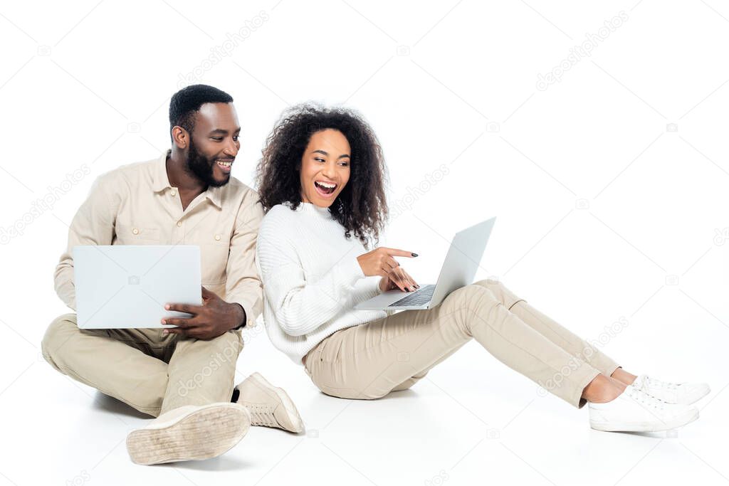 amazed african american woman pointing at laptop near smiling husband on white