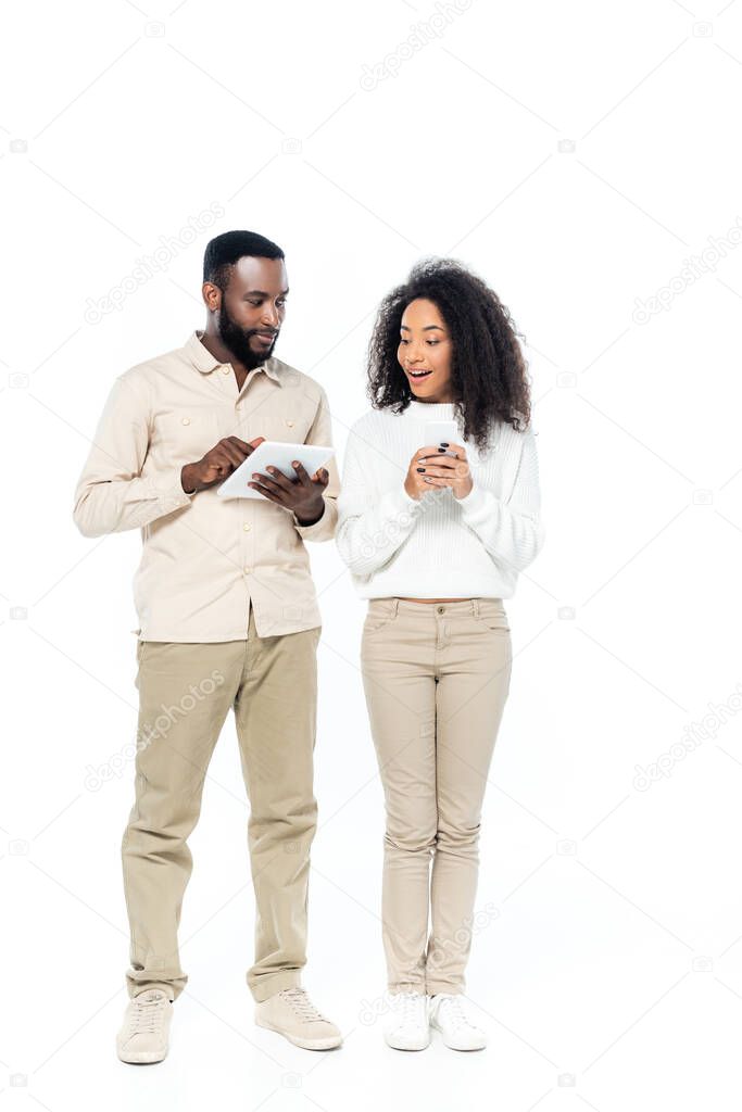 surprised african american woman holding smartphone while looking at digital tablet in hands of husband on white