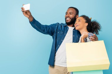 Mutlu Afro-Amerikalı adam, neşeli karısıyla selfie çekiyor. Elinde mavi torbalarla.